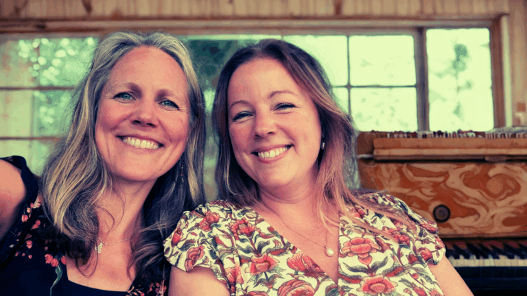 Twee lachende vrouwen poseren samen voor de camera in een kamer met grote ramen en een houten piano op de achtergrond.