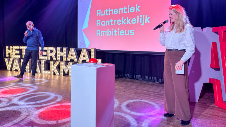 Twee mensen staan op een podium met microfoons; op de achtergrond zijn verlichte letters en een groot scherm met de woorden 'Authentiek', 'Aantrekkelijk', 'Ambitieus' te zien.