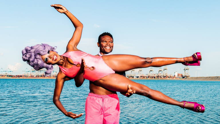 Twee personen bij het water; een draagt een roze badpak en paarse pruik en wordt in een luchtige pose gehouden door de ander die roze shorts draagt.