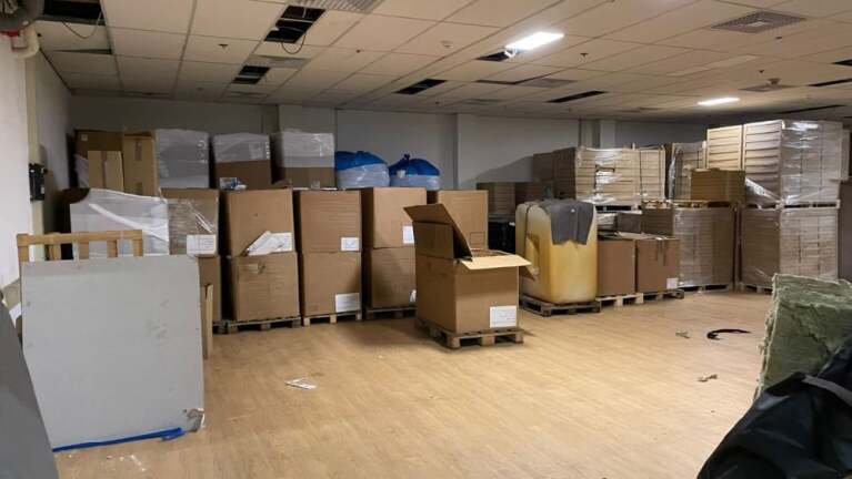 Opslagruimte gevuld met grote kartonnen dozen op pallets, sommige dozen zijn geopend. De ruimte heeft een systeemplafond met enkele ontbrekende platen en een houten vloer.