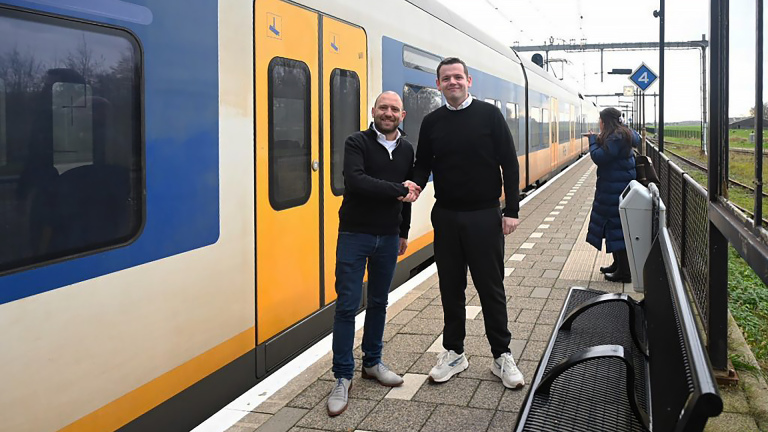 Twee mannen schudden elkaar de hand op een perron met een gele en blauwe trein ernaast, terwijl een vrouw op de achtergrond op haar telefoon kijkt.