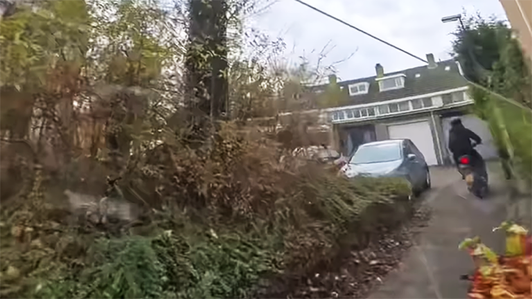 Een persoon op een motor rijdt langs een smalle oprit met een auto en een gebouw met garagepoorten aan de rechterzijde, omringd door herfstige bomen en struiken aan de linkerzijde.