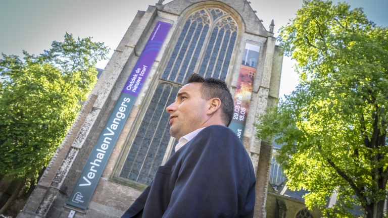 Een persoon in een pak staat voor een imposant kerkgebouw met grote glas-in-loodramen en banners met tekst, omringd door groene bomen.