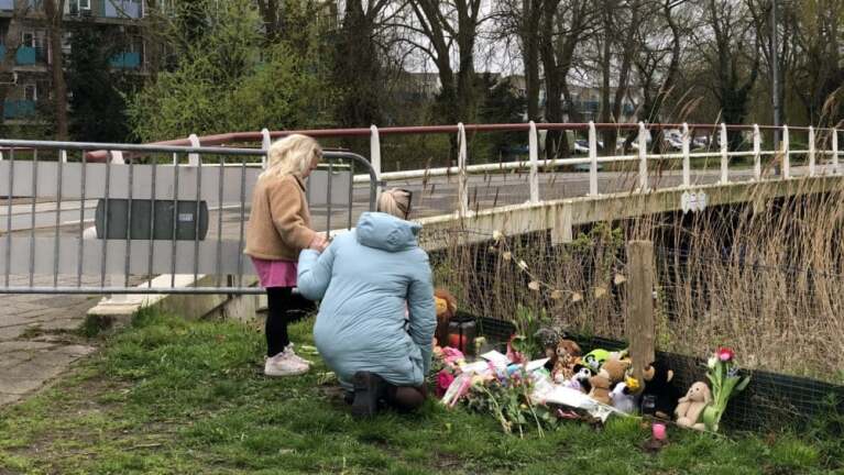 Twee personen, een volwassene en een kind, kijken naar een herdenkingsplaats naast een brug, versierd met bloemen en knuffels.