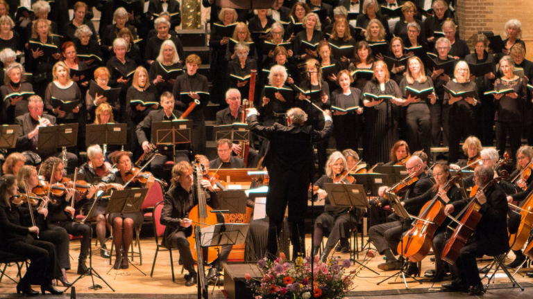 Een orkest speelt op een podium met een groot koor op de achtergrond, gedirigeerd door een man in een zwart pak, met verschillende strijkinstrumenten, blaasinstrumenten en zangers in formatie.