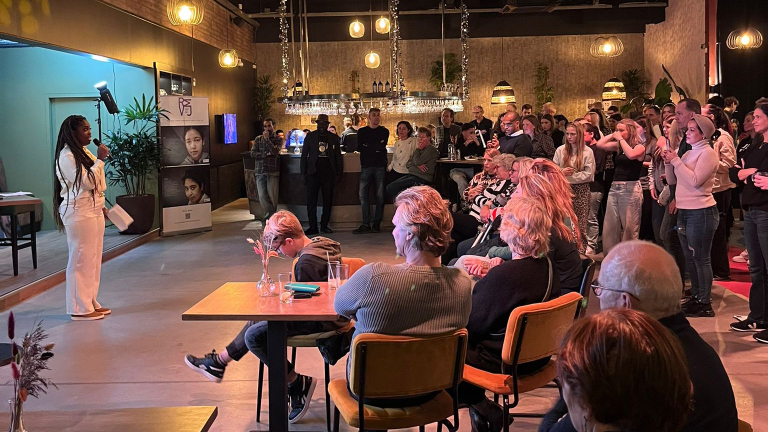 Een evenement waar een vrouw in het wit een toespraak houdt voor een publiek dat zit en staat, in een sfeervol verlichte ruimte met een bar op de achtergrond.