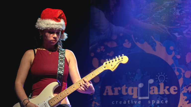 Vrouw speelt elektrische gitaar en draagt een kerstmuts, met achtergrondverlichting en een kunstzinnig logo op een blauw decor.