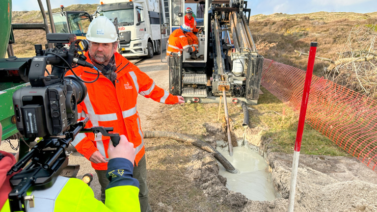 Man in een oranje veiligheidsjas en witte helm wordt gefilmd terwijl hij naast graafapparatuur en een werkput staat. Achter hem zijn twee andere arbeiders bezig met de machine, en op de achtergrond staan vrachtwagens. Er is een rode veiligheidshekken aan de zijkant van de werkplek.