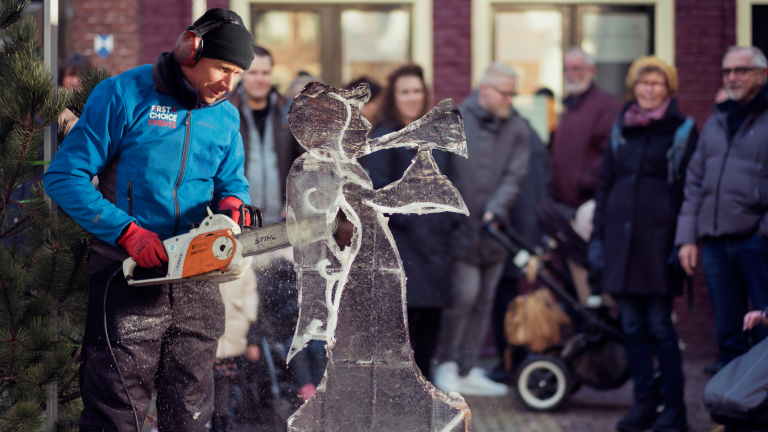 Een man in een blauw jack met de tekst 'First Choice Events' snijdt met een kettingzaag aan een ijssculptuur van een figuur met een trompet, terwijl een groep mensen toekijkt.
