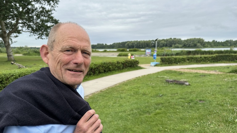 Ouder persoon in een parkachtige omgeving met een trui over de schouders en een lach op het gezicht, met gras, een pad, en bomen op de achtergrond.