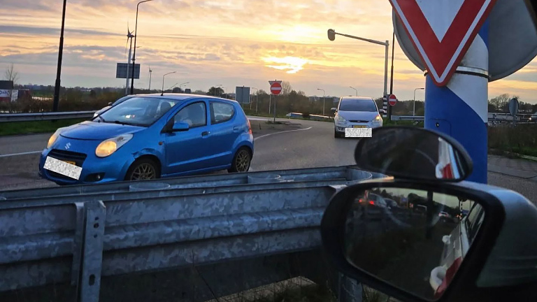Een blauwe auto rijdt op een snelwegafslag bij zonsopgang, met verkeersborden en een vangrail op de voorgrond.