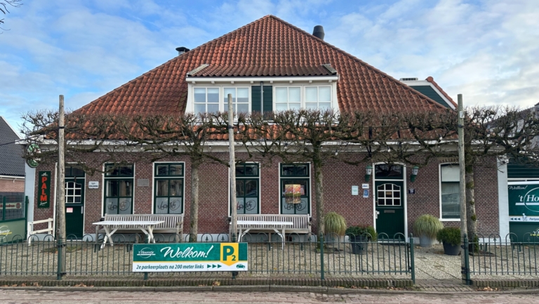 Een bakstenen gebouw met een rood pannendak en een opvallend bord met "Welkom!" dat een parkeerplaats aanwijst. Er zijn bomen met gesnoeide takken voor het gebouw en een hek met twee witte banken ervoor. De lucht is bewolkt met enkele blauwe plekken.