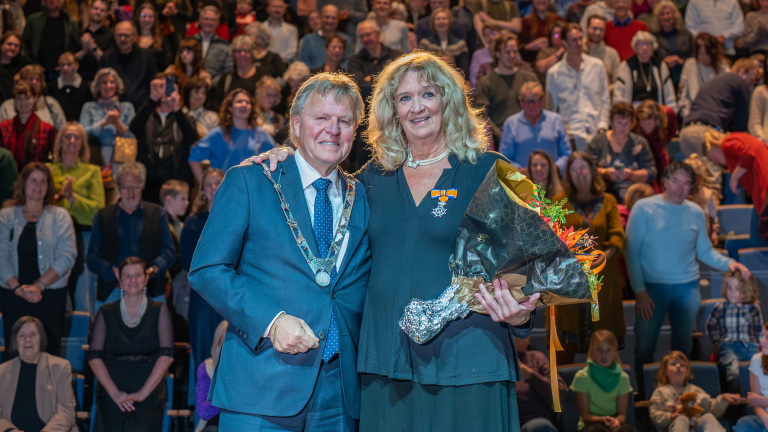 Twee personen poseren voor een volle zaal; een man in een blauw pak met ambtsketen en een vrouw met een bloemenboeket en onderscheidingslint op haar kleding.