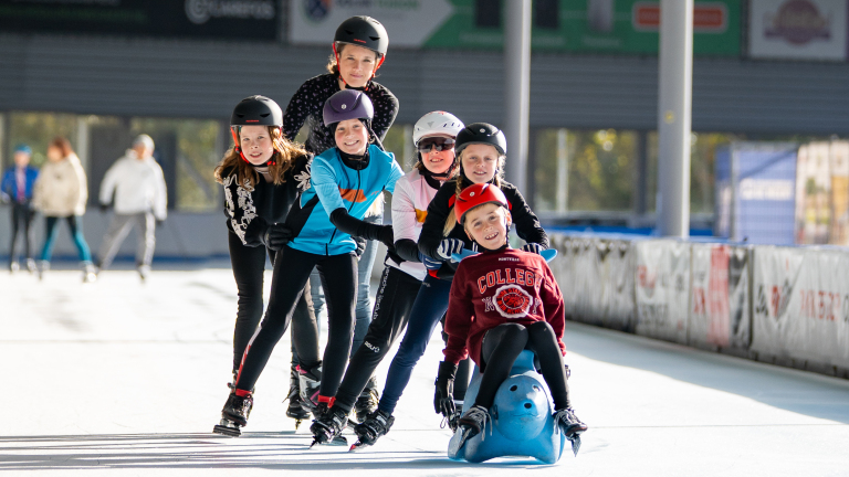 Groep kinderen in winterkleding en helmen schaatst in een rij op een ijsbaan, met een kind dat voorop zit op een blauwe slee. Ze lachen en hebben plezier.