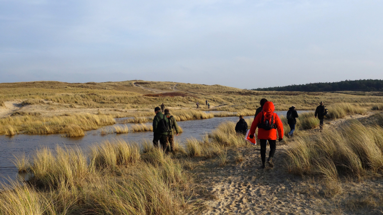 Een groep mensen wandelt door een duinlandschap met gras en een klein stroompje, onder een heldere hemel.