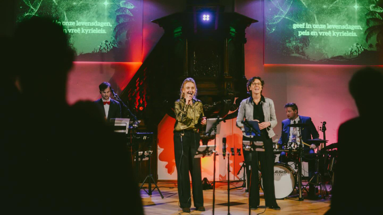 Een band treedt op op een podium, met een zangeres die in een microfoon zingt, een toetsenist, en een drummer op de achtergrond. Er is kleurrijke verlichting en een scherm met tekst in het Nederlands zichtbaar.