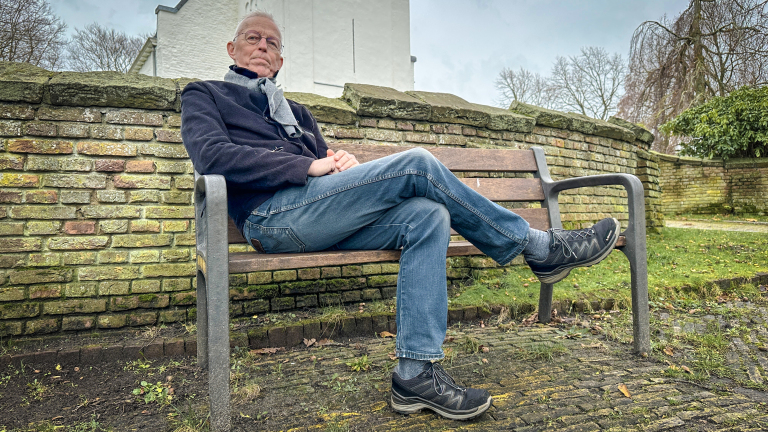 Een oudere man zit met gekruiste benen op een houten bankje in een park voor een stenen muur, omringd door gras en bomen.