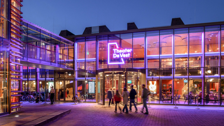 De ingang van Theater De Vest verlicht in paars licht, met mensen die het gebouw betreden, gefotografeerd in de schemering.