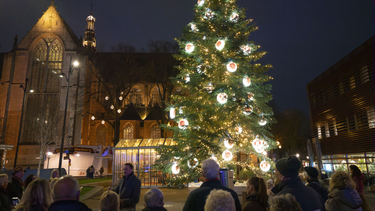Verlichte kerstboom met versieringen voor een verlichte historische kerk, omringd door toeschouwers in de avond.