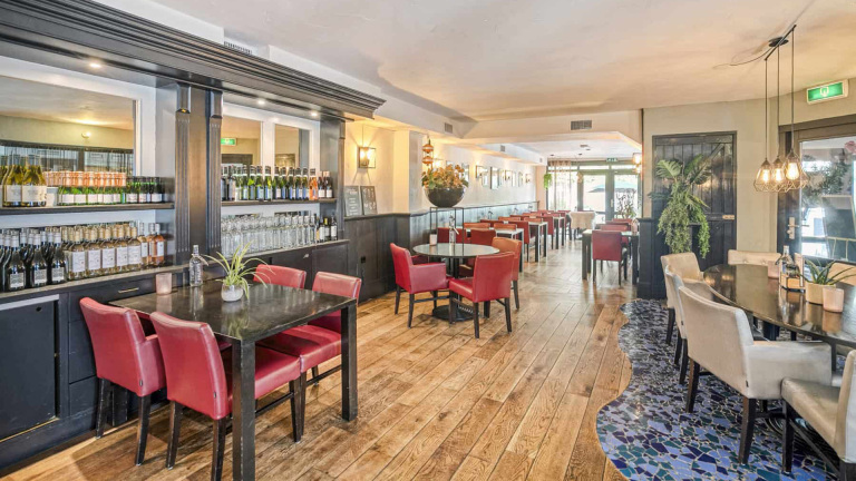 Lichte, ruime eetkamer van een restaurant met houten vloer, rode en witte gestoffeerde stoelen en tafels. Links is een wand met planken vol flessen wijn te zien, terwijl aan de rechterkant een decoratieve mozaïekvloer en planten zijn geplaatst. De ruimte is sfeervol verlicht en heeft een moderne uitstraling.