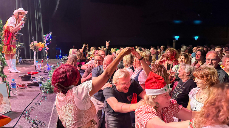Een zangeres in een opvallende outfit op een podium, omringd door een enthousiast publiek dat samen danst en lacht.