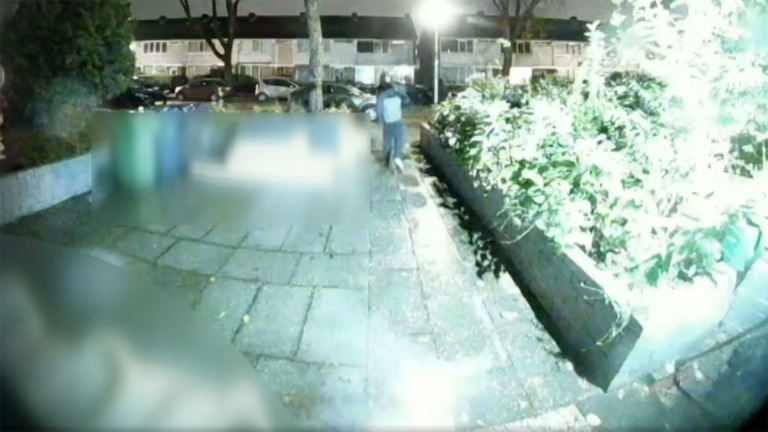 Nachtzicht van een straat met plantenbakken en een persoon die verderop over de stoep loopt, met huizen op de achtergrond.