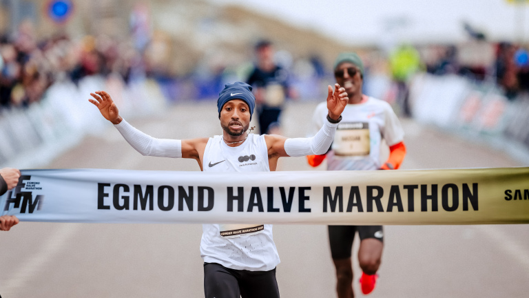 Hardloper passeert de finish van de Egmond Halve Marathon, met armen omhoog, voor een juichend publiek.