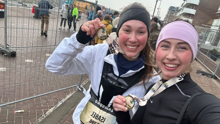 Twee lachende vrouwen met medailles na een marathon, omringd door andere lopers en hekken.