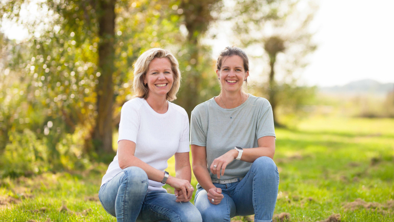 Twee vrouwen zitten op een grasveld in een zonnig park, glimlachend naar de camera.