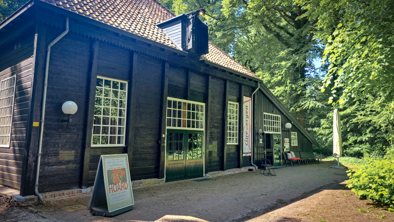 Houten gebouw met grote ramen en een bruin pannendak, omgeven door groen op een zonnige dag, met een bord waarop 'Hoard' staat, voor de ingang.