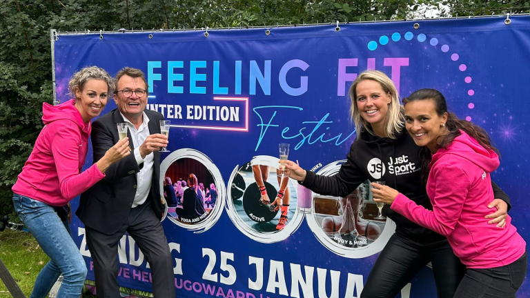 Vier mensen poseren glimlachend voor een spandoek met de tekst "Feeling Fit Winter Edition" en houden glazen vast.