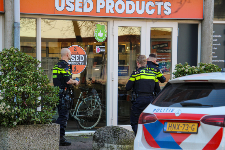 Agenten onderzoeken een winkel met "Used Products" op de gevel, naast een geparkeerde politiewagen.