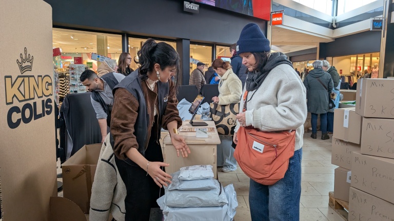 Mensen staan in een winkelcentrum bij een kraam van "King Colis" waar pakketten worden uitgestald en besproken.