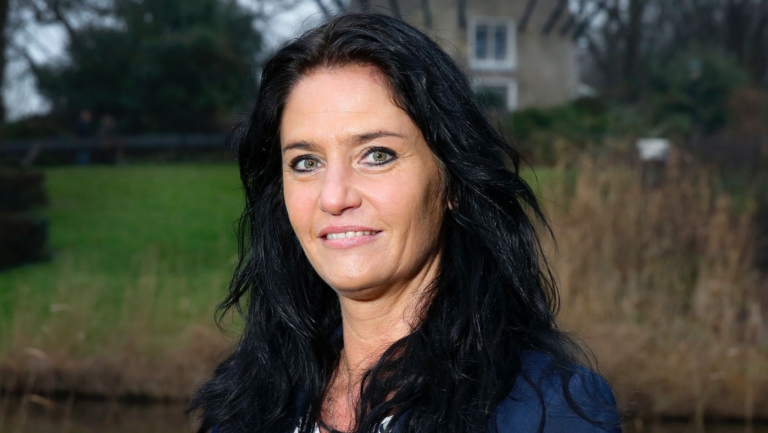 Vrouw met donker haar en een blauwe blazer staat buiten met een molen op de achtergrond.