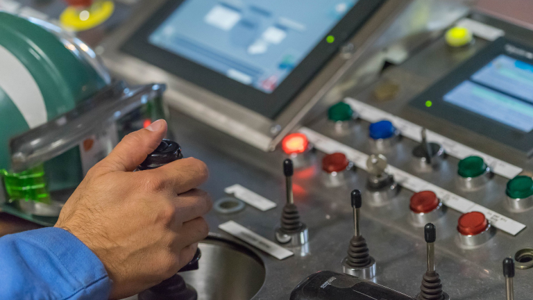 Hand bedient joystick op een controlepaneel met knoppen en schermen. Een groene helm ligt ernaast.