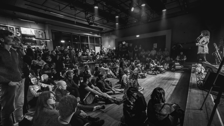 Een vrouw met microfoon spreekt voor een groot zittend publiek in een intieme theateromgeving.