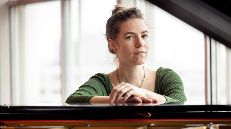 Vrouw in groene blouse leunt op een piano, met ramen op de achtergrond.