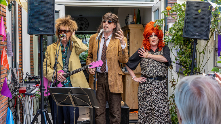 Optreden van drie personen in kleurrijke kostuums met microfoons, een gitaar en luidsprekers in een tuinsetting.