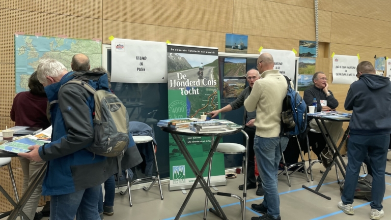 Mensen staan bij informatiestands over fietsroutes, waaronder "De Honderd Cols Tocht", met kaarten en brochures op hoge tafels.