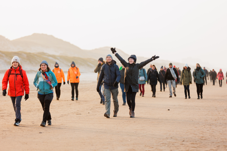 Een groep mensen wandelt over een strand, sommige dragen kleurrijke windjacks en mutsen, terwijl een vrouw haar armen wijd uitstrekt.