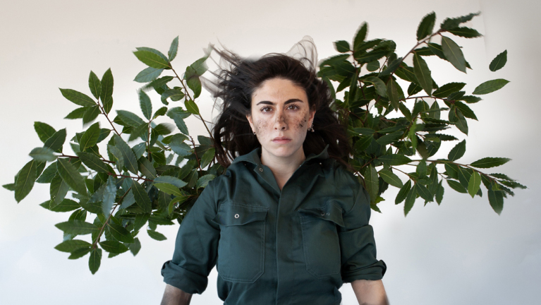 Een vrouw met een groene blouse staat voor een achtergrond van groene bladeren, met wapperend haar en vlekken op haar gezicht.