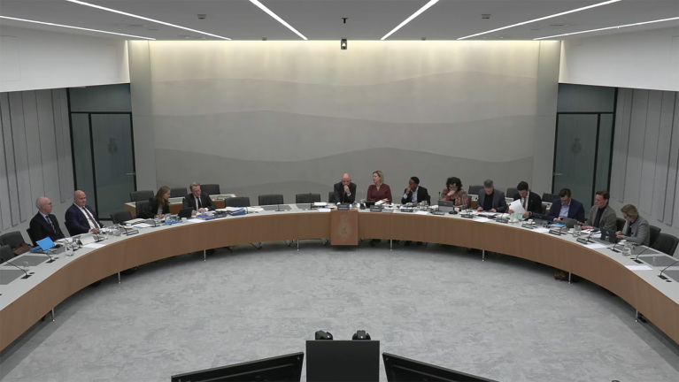 Mensen zitten aan een grote, gebogen tafel in een vergaderruimte, bezig met documentatie en laptops.