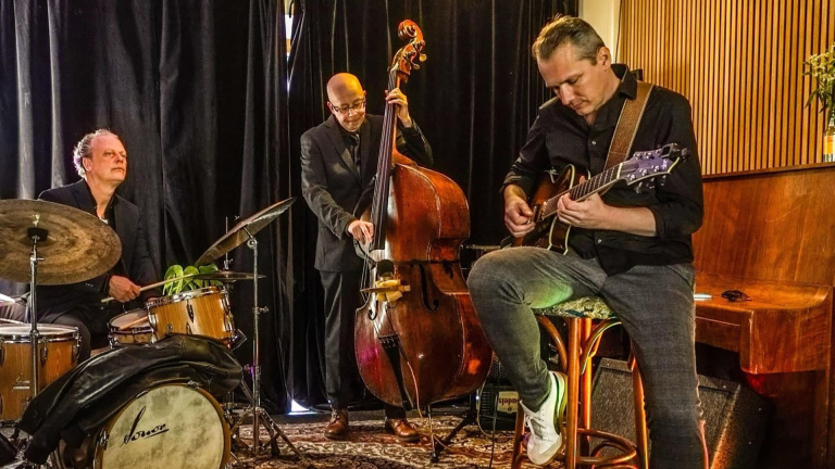 Drie muzikanten spelen samen; een drummer, een contrabassist en een gitarist in een kamer met een donkere gordijnachtergrond.