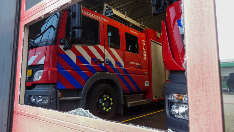 Rode brandweerwagen met blauwe en witte strepen in een garage, gezien door een gebroken raampaneel.