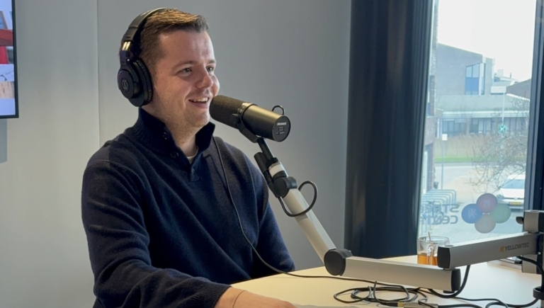 Man met koptelefoon en microfoon in opnamestudio, met gele dobbelsteen op tafel.