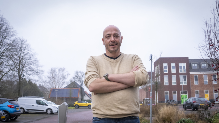Man in een beige trui met gekruiste armen staat buiten voor een gebouw en geparkeerde auto's.