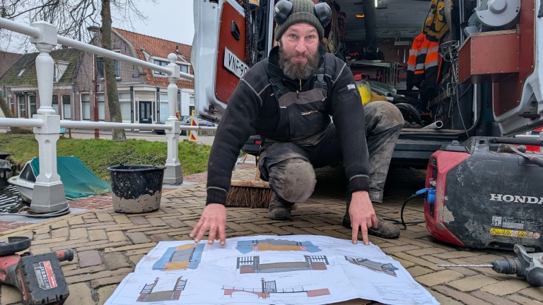 Man met werkpak bestudeert een bouwtekening op de grond naast een open busje en gereedschap bij een kanaal.