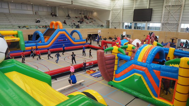 Kinderen spelen in een sporthal met opblaasbare attracties en hindernisbanen, terwijl toeschouwers vanaf de tribune toekijken.