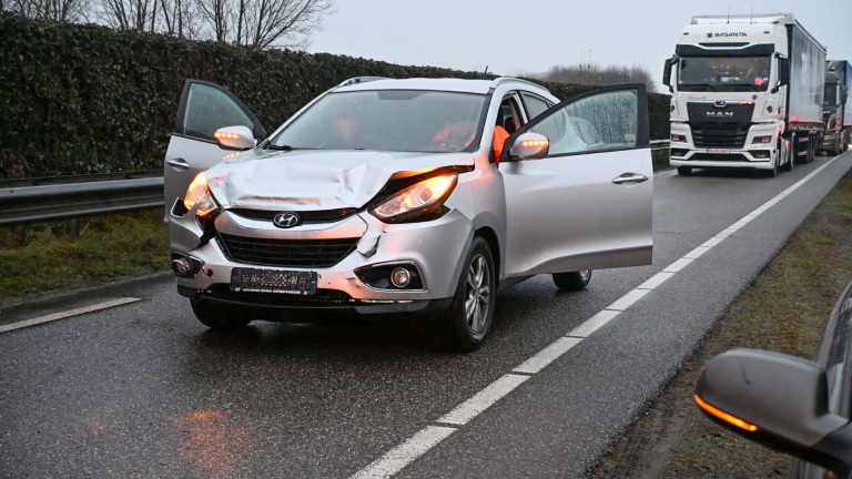 Auto met schade aan de voorkant op een weg, met open portieren en een vrachtwagen op de achtergrond.