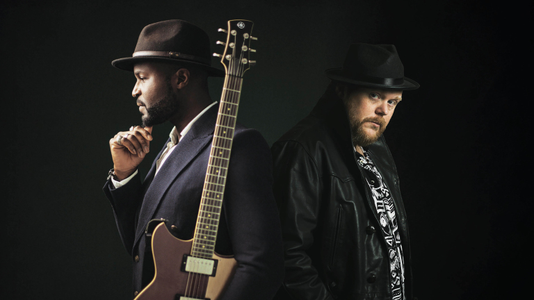 Twee mannen met hoeden; één met gitaar, kijkt opzij, de ander draagt een leren jas en kijkt naar de camera.